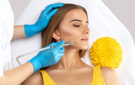 Photo for A cosmetologist doctor performs a microdermabrasion procedure on the face against acne and blackheads in a beautiful young girl in yellow. Women's cosmetology in a beauty salon. - Royalty Free Image