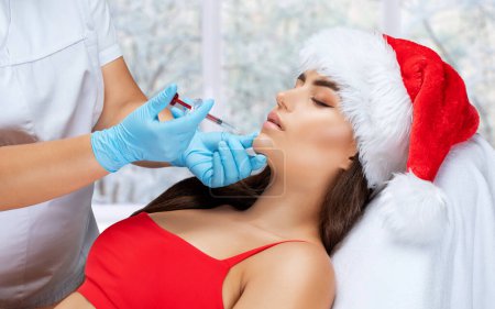 El cosmetólogo hace anti-envejecimiento inyecciones anti-arrugas en la cara de una hermosa mujer en un sombrero de santa rojo. Cosmetología estética femenina en un salón de belleza. Concepto de Año Nuevo.