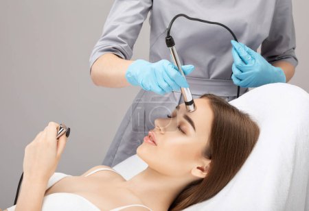 A doctor cosmetologist makes a microcurrent facial therapy to a young woman with a device in a beauty wellness salon.Cosmetology and professional skin care.