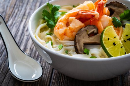 Tom Kha goong soup - coconut shrimp soup with noodles and shiitake mushrooms on wooden table