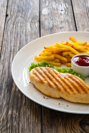 Grilled chicken breast with fries and fresh vegetables on wooden table 