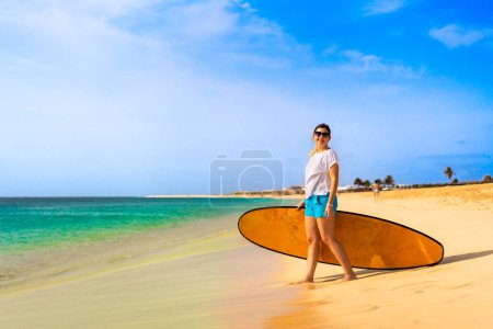 Beautiful woman holding surfboard standing on sunny beach Santa Maria, Sal island , Cape Verde  Poster 704223322