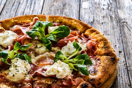 Circle prosciutto pizza with mascarpone cheese and leafy greens on wooden table 