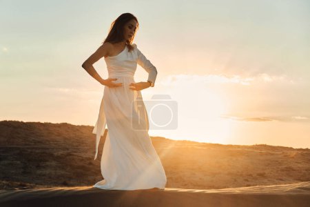 Téléchargez les photos : Une fille en robe blanche danse et pose dans le désert de sable au coucher du soleil. - en image libre de droit
