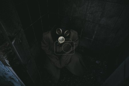 Foto de Un hombre con una máscara de gas se sienta en una pequeña habitación oscura en sus haunches y mira hacia arriba, buena cubierta de libro fo - Imagen libre de derechos