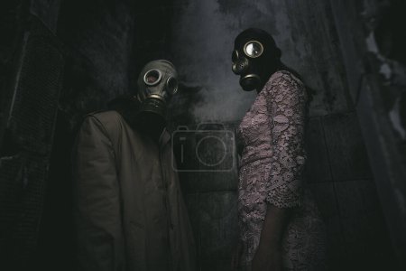 Photo for Two people in gas masks stand against a wall in a small dark room, low angle shot , good for book cover - Royalty Free Image