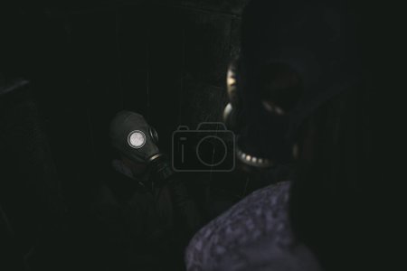 Photo for A girl in a gas mask is viewed from the back, standing and looking down at a sitting guy in a gas mask, in a dark small room. Good for book cover - Royalty Free Image