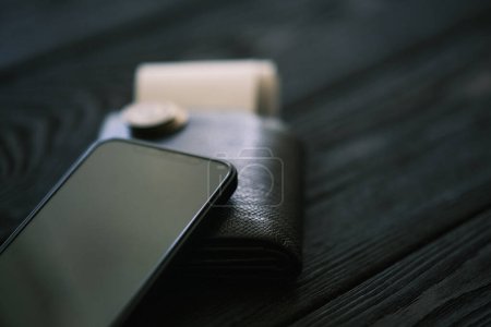 Foto de Un teléfono móvil que pone en un bolso de cuero y pila de monedas en la mesa de madera negro, concepto financiero - Imagen libre de derechos