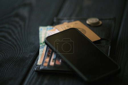 Foto de Un teléfono móvil que pone en un bolso de cuero y una pequeña pila de monedas en la mesa de madera negro, concepto financiero - Imagen libre de derechos