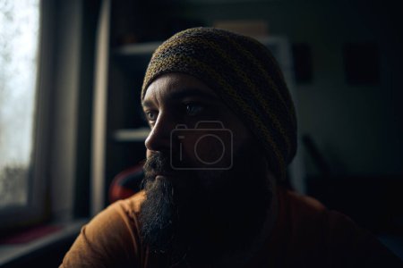 Téléchargez les photos : Un homme barbu portant un chapeau rayé et un t-shirt, regardant par la fenêtre - en image libre de droit