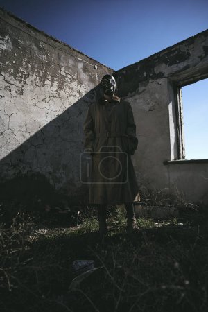 Foto de Una persona de pie frente a una pared con una máscara de gas y gabardina, en la hierba, tiro de ángulo bajo, bueno para la cubierta del libro - Imagen libre de derechos