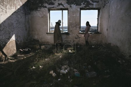 Photo for Two people, a guy and a girl in gas masks, stand inside an abandoned house next to the window and look back, apocalypse - Royalty Free Image