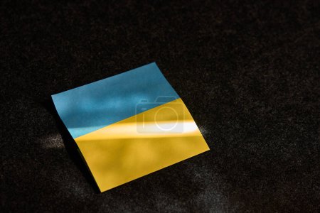 Téléchargez les photos : L'autocollant sous la forme du drapeau ukrainien se trouve. sur une surface sombre et un rayon de soleil tombe dessus - en image libre de droit