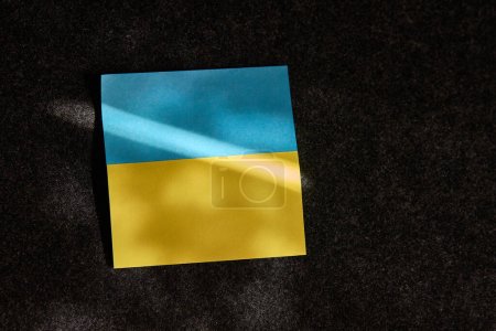 Foto de La pegatina en forma de bandera ucraniana se encuentra. en una superficie oscura y un rayo de luz solar cae sobre ella - Imagen libre de derechos