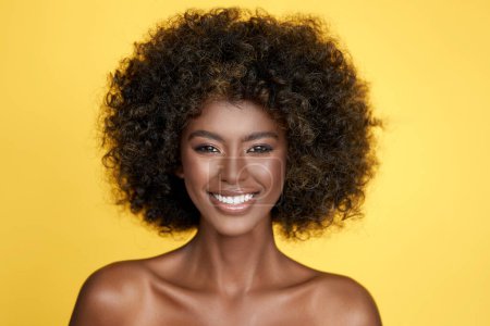 Optimistic curly haired female model with bare shoulders looking at camera isolated on yellow background