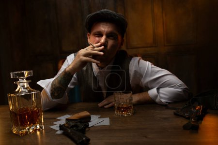 Cinematic portrait of young 1920s British gangster in waistcoat and flat cap sitting at table with cards and gun lying nearby in vintage casino, smoking and drinking whiskey. Peaky blinders style look