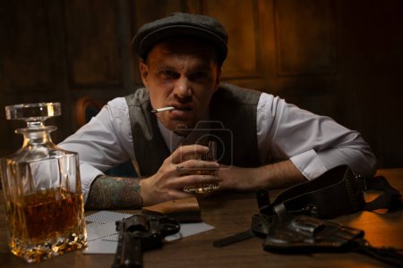 Aggressive young gangster sitting with cigarette in mouth and glass of whiskey in hands in vintage casino, looking frowning at camera. Cards, revolver and holster lying in front of man on table