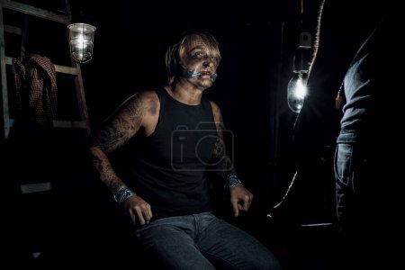 Foto de Hombre adulto con tatuajes en brazos y rostro y cuerpo magullados, secuestrado, sentado amordazado con las manos atadas a la silla en el sótano oscuro, mirando con miedo a su captor. Secuestro, violencia y delincuencia - Imagen libre de derechos