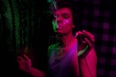 Téléchargez les photos : Portrait d'un adolescent aux cheveux foncés lapidé avec des paillettes brillantes sur les joues debout dans une pièce sombre avec cigarette à la main, regardant avec défiance la caméra et soufflant de la fumée de sa bouche - en image libre de droit