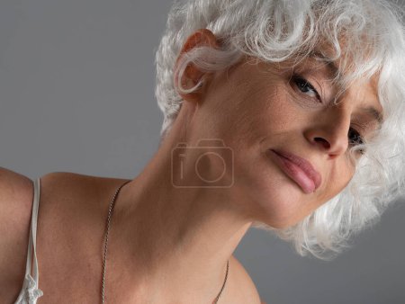 Closeup portrait of confident attractive senior woman with wavy white hair falling over face posing in studio on gray background, looking at camera