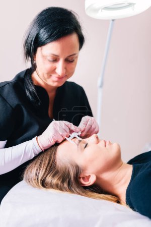 Beautician measuring for eyebrow geometry, shaping or lamination cosmetic procedure on woman's face in a beauty salon,