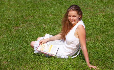 Photo for Happy female student sitting on the grass with a book - Royalty Free Image