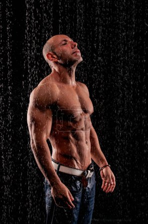 Téléchargez les photos : Bodybuilder athlète sous des jets de pluie sur un fond noir - en image libre de droit