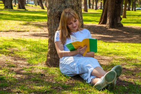 Foto de Una chica con un libro se sienta cerca de un árbol en el parque - Imagen libre de derechos