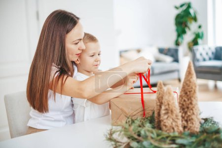 Foto de Madre y su hijo pequeño empacando una gran caja de regalo de Navidad juntos en casa, atando un lazo en un gran regalo de Navidad - Imagen libre de derechos