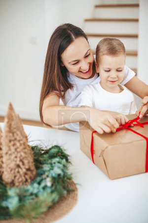 Foto de Sonriente madre y su pequeño hijo empacando una gran caja de regalo de Navidad juntos en casa, atando un lazo en el presente - Imagen libre de derechos