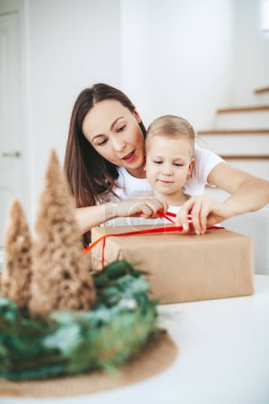 Foto de Madre y su pequeño hijo atando un lazo en una gran caja de regalo de Navidad juntos en casa, embalaje de regalos de Navidad - Imagen libre de derechos