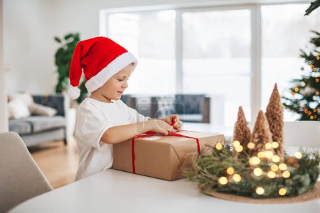 Photo for Cute little boy in red Santa Claus hat untying a bow on a big gift box. - Royalty Free Image