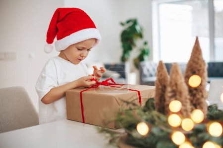 Photo for Serious little boy in red Santa Claus hat untying a bow on a big gift box. Merry Christmas and Happy New Year concept. - Royalty Free Image