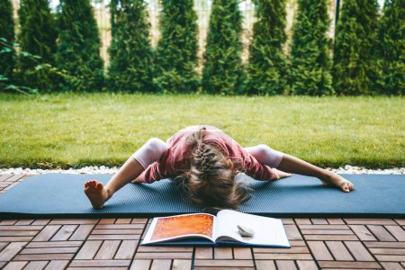 Photo for Little girl 5-6 years old lying in the Turtle pose or Kurmasana while practicing yoga outdoors. Yoga for children. Kid's healthy lifestyle. - Royalty Free Image