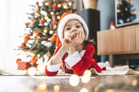 Linda niña de 5-6 años con sombrero de Santa escribiendo sus deseos a Santa por el árbol festivo, sus ojos llenos de sueños