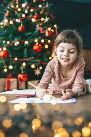 Niña de 5-6 años escribiendo su carta a Santa cerca del árbol de Año Nuevo bellamente decorado