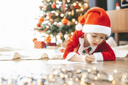 Cute little girl 5-6 years old by the holiday tree, putting her hopes and dreams into a letter for Santa Claus
