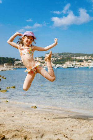 Foto de Niña feliz 5-6 años de edad en un traje de baño saltando en la playa, exudando pura alegría y emoción en medio del telón de fondo costero arenoso - Imagen libre de derechos