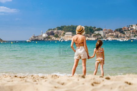 Frau und ihre kleine Tochter im Badeanzug stehen zusammen am Strand und beobachten die Bucht von Port de Soller auf Mallorca. Ferien mit Kindern.