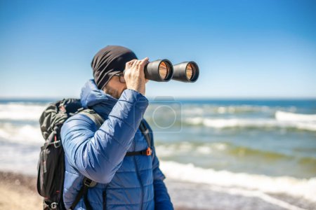 Foto de Un turista de pie junto a la orilla del mar, mirando a través de prismáticos, realizando una exploración visual del entorno costero - Imagen libre de derechos