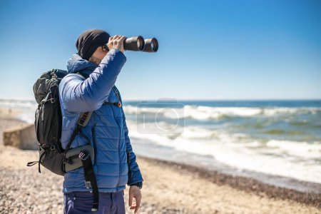 Foto de Un turista, parado a la orilla del mar, usando prismáticos para ver más de cerca el paisaje costero - Imagen libre de derechos