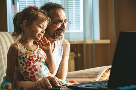 Cute little girl 5-6 years old and her grandfather 60-65 years old engaged with a laptop. Two generations learning how to use technologies.