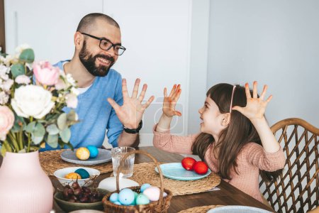 Cute little girl and her father playing with red Easter eggs at home. Celebrating and eating Easter breakfast concept.