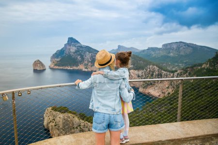 Una mujer con sombrero de paja abrazando a su hijita, mientras contempla una vista impresionante desde el Mirador de El Colomer en Mallorca. Viajar con concepto de niños.
