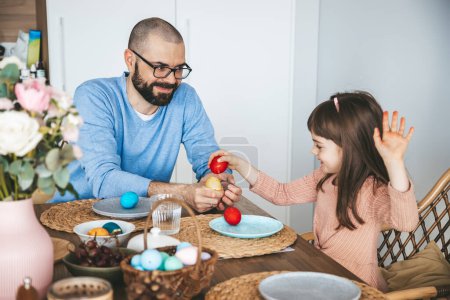 Little girl and her father playing an egg fight game with red Easter eggs, sitting at the dining table. Celebrating and eating Easter breakfast concept.