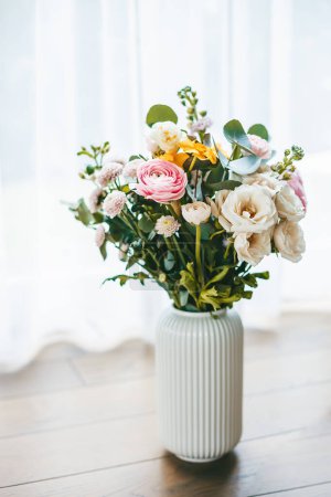 Un bouquet vibrant de fleurs en différentes couleurs et types, disposées dans un vase blanc côtelé sur un sol en bois sur fond de fenêtre avec des rideaux transparents, illuminant la scène avec de la lumière naturelle