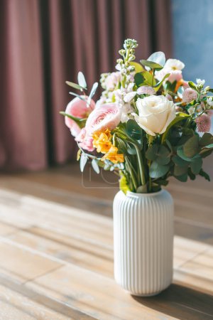 Téléchargez les photos : Le bouquet de fleurs assorties comprenant des roses blanches, des ranunculus roses et des fleurs jaunes, dans un vase côtelé blanc, placé sur un sol en bois sur fond de rideaux roses - en image libre de droit