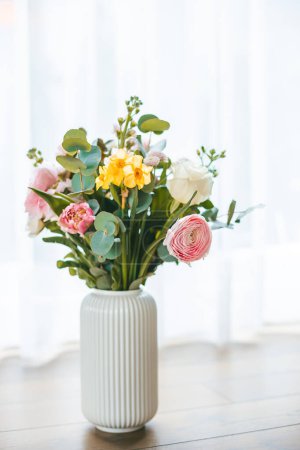 Un bouquet vibrant de fleurs éclate avec un éventail de couleurs et de types, tenu dans un vase blanc côtelé sur fond de fenêtre, baigné de lumière naturelle douce