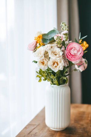 Ein bunter Blumenstrauß in verschiedenen Farben und Typen, arrangiert in einer weißen, gerippten Vase auf einer Holzoberfläche, vor dem Hintergrund eines Fensters mit weißen Vorhängen