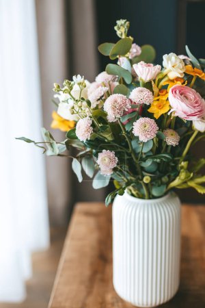 Un bouquet vibrant de diverses fleurs disposées dans un vase blanc côtelé qui s'assoit sur une table en bois, avec la lumière naturelle douce mettant en valeur les couleurs et les textures des fleurs